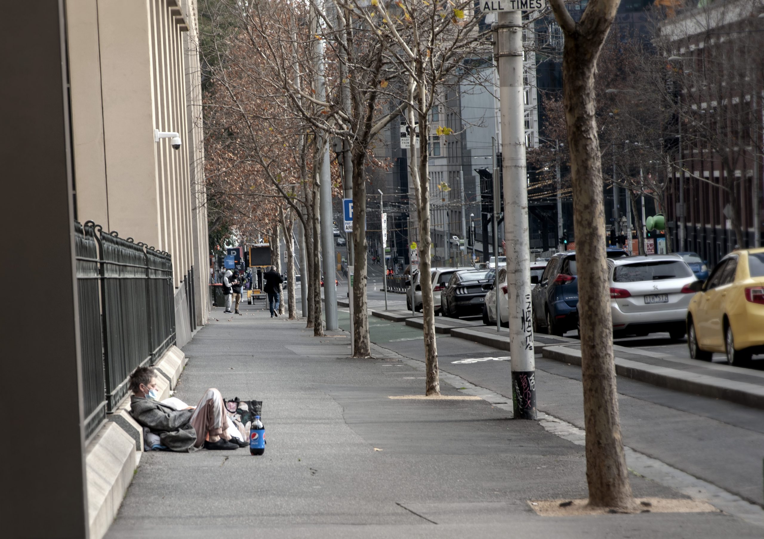 Bezdomny siedzący na ulicy, w maseczce na ustach. Ulica przy głównej bibliotece stanowej w Melbourne.